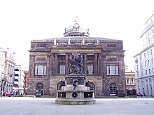 A Neoclassical building seen end-on. It has two storeys with a central colonnaded portico and the dome can be seen above the roof. In the foreground is an elaborate monument