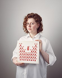 Rufina Bazlova ist stehend abgebildet. Sie hält ein quadratisches Kunstwerk, auf dem rote Piktogramme auf weißem Untergrund zu sehen sind. Bazlovas rotes lockiges Haar ist halblang und sie trägt eine weite weiße Bluse. Sie schaut direkt in die Kamera.