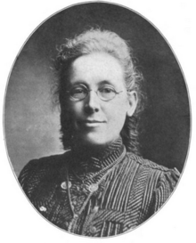 A middle-aged white woman, with grey hair dressed back away from her face. She is wearing glasses and a high-collared striped dress or blouse.