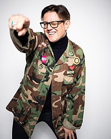 Portrait of Seven Graham, wearing a camouflage jacket and pointing at the camera and winking