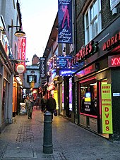 Narrow, pedestrian alleyway, with sex clubs, adult cinemas, strip clubs and a tattoo parlour all visible, all advertising their presence with neon signage.