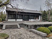 Traditional Chinese-style building with dark wooden beams and large windows, surrounded by a paved stone area and greenery.