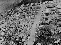 A mass of jumbled wreckage beside a street with pedestrians and vehicles nearby