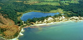 Aerial photograph of a lakeshore, a sandy barrier beach, and a pond behind it; the photograph appears to have been taken in summer, since the land around the pond is green with foliage. There are high, forested sand dunes on the barrier beach.