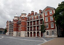 A four-story building of light grey stone and red brick with a complex facade including two central towers, and pillar and arch verandahs on the face of three of the four storeys