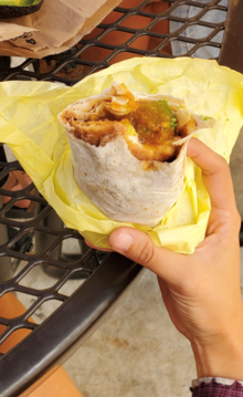 A photograph of a hand holding a chile relleno burrito wrapped in yellow paper in front of a brown metal picnic table.