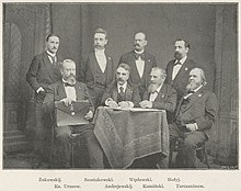 Black and white photo; seven men in suits sit and stand around a table