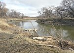 A small river flows through a prairie landscape; brown grasses and leafless trees line the banks.