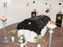 Small chamber with white walls and pink marble floor. In the middle is a tomb on three small stairs from the same marble, covered with white silk and black velvet with embroidered golden Greek crosses. It is surrounded by candles and flowers.