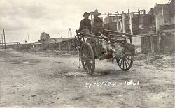 Two men in sombreros riding in a donkey-cart with a line of feet sticking out the back. They are riding down a dirt street away from the camera, with a line of buildings on the right. Dated 5/15/1911.