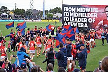 Pearce running towards a banner bearing her portrait followed by her teammates, with Melbourne AFL players and staff forming a guard of honour and fans visible in the background