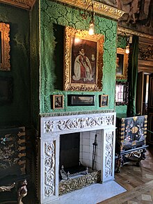 A photograph of a richly-decorated room, covered in bright green silk damask over which several small paintings are hanging. A white marble fireplace and two lacquer cabinets are also visible.