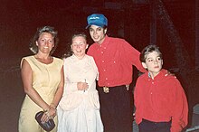 A smiling Jackson wears a blue baseball cap and a red shirt. On his right are two women. One holds a pen, and one a small purse. On his left a young boy looks off-camera. He is dressed in a red shirt too. Jackson's hand is on his shoulder.