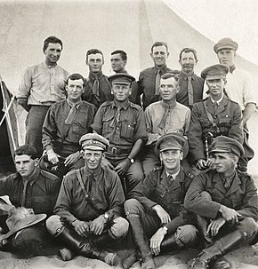 a group of officers in various forms of dress, some with headdress, sitting in front of a tent