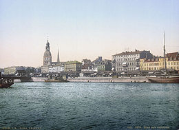 Alte Farbfotografie von einer Stadt mit der Aufschrift „Riga Le Quai“. Im Vordergrund ist ein breiter Fluss mit drei Schiffen. Am Ufer sind überdachte Stände und eine Brücke ist links teilweise zu sehen. Hinter der linken Häuserfassade sind zwei Kirchtürme.