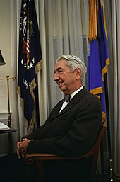 Photograph of a man in a suit, seated, with flags in the background