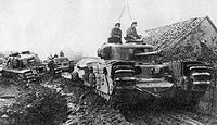 tanks moving along a muddy road beside a low building