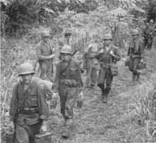 Black and white photo of four men wearing military uniforms and helmets carrying bags and boxes along a trail. Several other men are standing at the edge of the trail, and behind them
