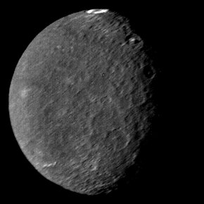 A round spherical body with its left half illuminated. The surface is dark and has a low contrast. There are only a few bright patches. The terminator is slightly to the right from the center and runs from the top to bottom. A large crater named Wunda with a bright ring on its floor can be seen at the top of the image near the terminator. A pair of large craters with bright central peaks can be seen along the terminator in the upper part of the body. The illuminated surface is covered by a large number of craters.