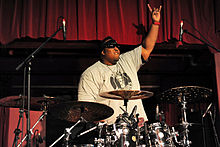 The drummer from the band Suicidal Tendencies, Eric Moore, is shown behind his drumkit. One hand is raised with the index finger and pinky extended.