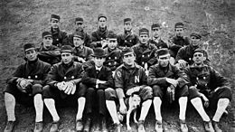 A black and white photograph of seventeen men sitting in three rows on the ground. Most are wearing dark coats with high white socks, but two are wearing dark baseball uniforms with a white "N" on the chests; some are wearing fielding gloves, and one is petting a dog.