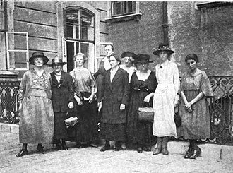 Nine women in dresses standing in front of a wrought iron fence behind which is a building with two open windows