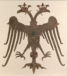 Photograph of a golden embroidered-double-headed eagle on an off-white background, with crowned heads and spread wings and legs, carrying a round medallion with Greek inscriptions on its breast.