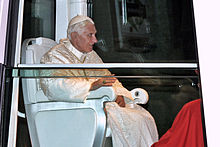 Seitliche Farbfotografie vom Papst in einem geschlossenen Fahrzeug. Er sitzt in einem weißen Stuhl und trägt helle liturgische Kleidung mit Kappe.