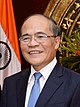 a smiling nearly bald man, wearing glasses, a suit and a blue tie, back him is flag of India
