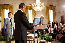 Obama in blue suit standing at a podium in front of an audience as a man in a light grey suit looks on.