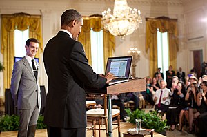 Black man in blue suit standing at a podium in front of an audience as a white man in a light grey suit looks on.