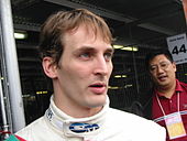 A man in his mid-twenties in white and turquoise racing overalls looking to the left of the camera