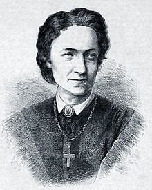 A middle-aged woman with shoulder-length black hair looking to her left dressed in a high-necked shirt and a large cross on a chain