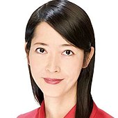 Aya Kamikawa ist in die Kamera schauend porträtiert. Sie hat langes glatte schwarzes, rechts gescheiteltes Haar. Ihre Lippen sind dezent rot geschminkt und sie lächelt leicht. Sie hat dunkle Augen und ein rotes Oberteil ist zu sehen.