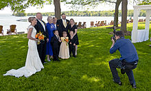 A man in a blue shirt and dark pants at right crouches as he takes a picture of a group of people at left in formal wear, with a woman in a white wedding dress at the rightmost. Behind them are tall trees and a large lake