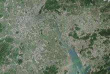 Satellite imagery showing a large grey coastal urban area with patches of green vegetation.