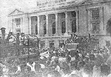 A black-and-white photograph of soldiers in the backs of army trucks surrounded by civilians in front of the National Palace of El Salvador