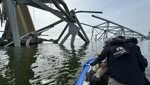 Three people in FBI uniforms are on a boat. They are closely inspecting the mangled remains of bridge struts poking out of the water in front of them.