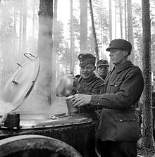 During additional refresher training, a Finnish soldier has his breakfast served into a mess kit by another soldier from a steaming field kitchen in the forests of the Karelian Isthmus. More soldiers, two of them visible, wait in line for their turn behind him. It is early October, and the snow has not yet set in.