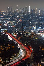 Aerial photograph of a freeway at night with car traffic turned into light trails with a long exposure; the skyline of Downtown Los Angeles is seen in the background.