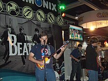 A color photograph of three young men playing on the The Beatles: Rock Band instruments in front of a large display for the game