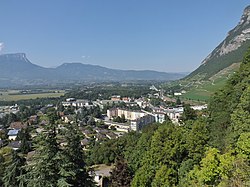 Photo shows homes and apartments in the center and a mountainside to the right. In the background are more mountains.