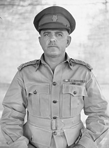 a male officer with a moustache wearing a peaked cap, khaki uniform and medal ribbons