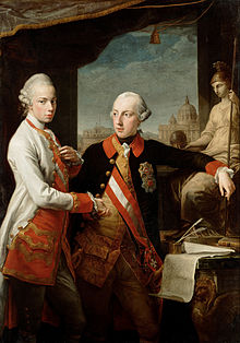 Two young men with hair powdered white, one in white-and-red, the other in black-and-gold military uniforms standing next to a statue of a Greek or Roman goddess