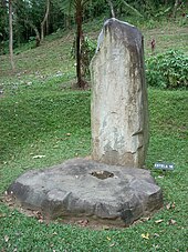A tall plain standing stone behind a cog-shaped flat stone, in a grassy area with a jungle-covered mound rising behind.