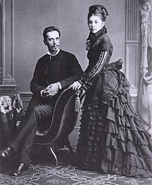 A lavishly dressed couple posing; the man on the left is sitting and the woman on the right stands next to him
