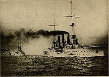 A large warship leaving a trail of thick, black smoke steams in line with three other vessels following in the distance.