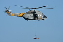 Spanish Air Force Aérospatiale SA330J Puma of 801 Squadron flying in an airshow. It is lifting a stretcher with a hoist. On the side of the helicopter is lettering reading "SAR", in yellow against the military grey colour scheme.