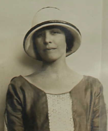 A young white woman with dark hair, wearing a cloche-style light-colored hat and a loose dress with a scooped neckline