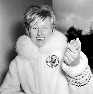 Anne Heggtveit wearing a plush coat, holds up a round medal.